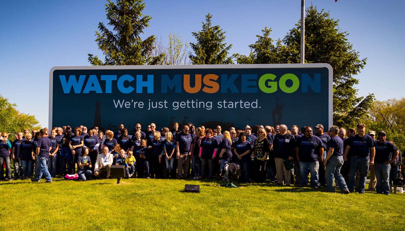 A group of people standing in front of a sign that says watch muskegon.